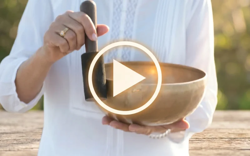 rosanaturae demostrando la técnica para tocar un cuenco tibetano artesanal de siete metales con mazo de madera, imagen con botón de reproducción para tutorial de sonoterapia.